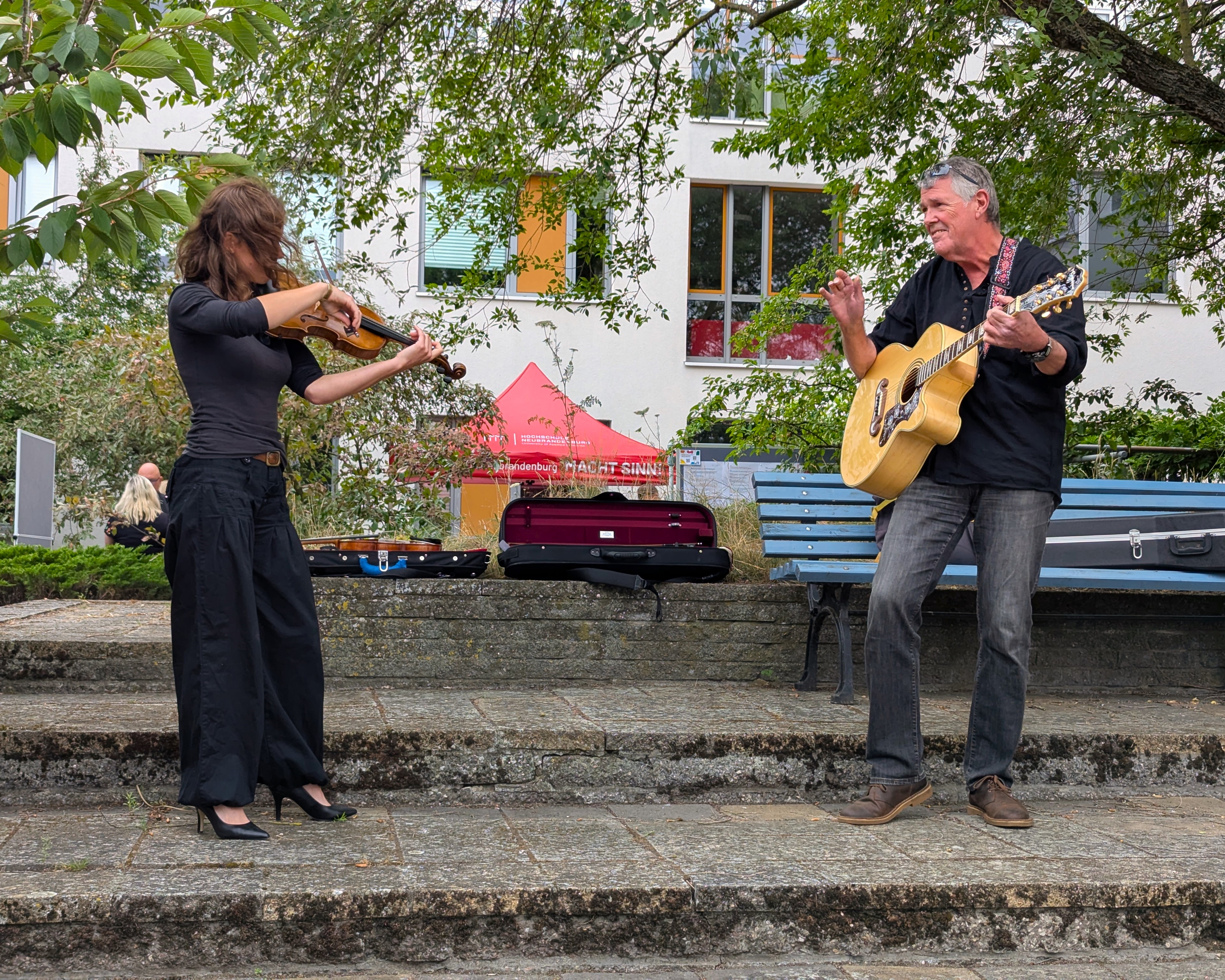 Zwei Personen stehen im Innenhof und musizieren. Die Person im linken Bildrand spielt Violine. Ihre braunen langen Haare wehen ihr ins Gesicht. Die zweite Person steht rechts von ihr. Der Mann spielt Akustikgitarre und sieht freundlich in die Richtung der Violinistin.