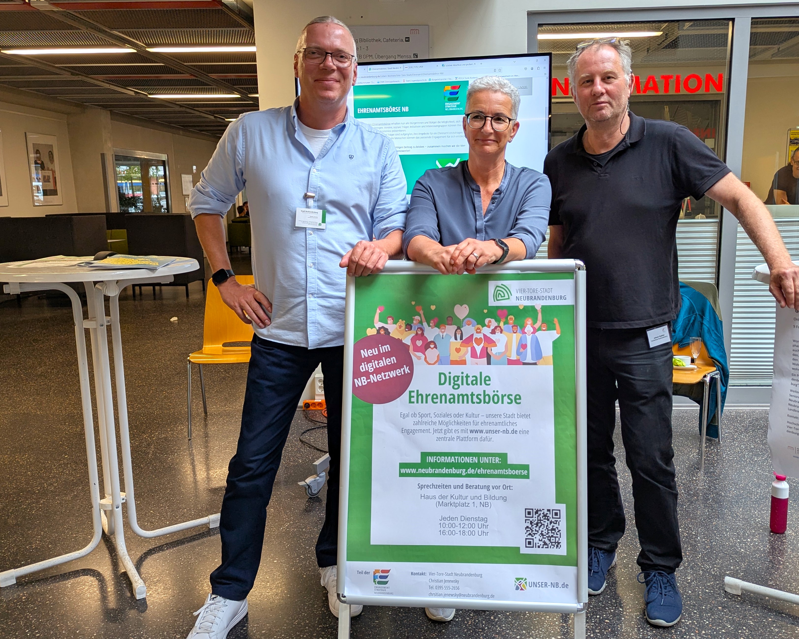 Auf dem Foto sind drei Personen zu erkennen, die freundlich in die Kamera schauen. Vor ihnen steht ein Aufsteller mit einem Hinweis auf die Digitale Ehrenamtsbörse der Stadt Neubrandenburg.