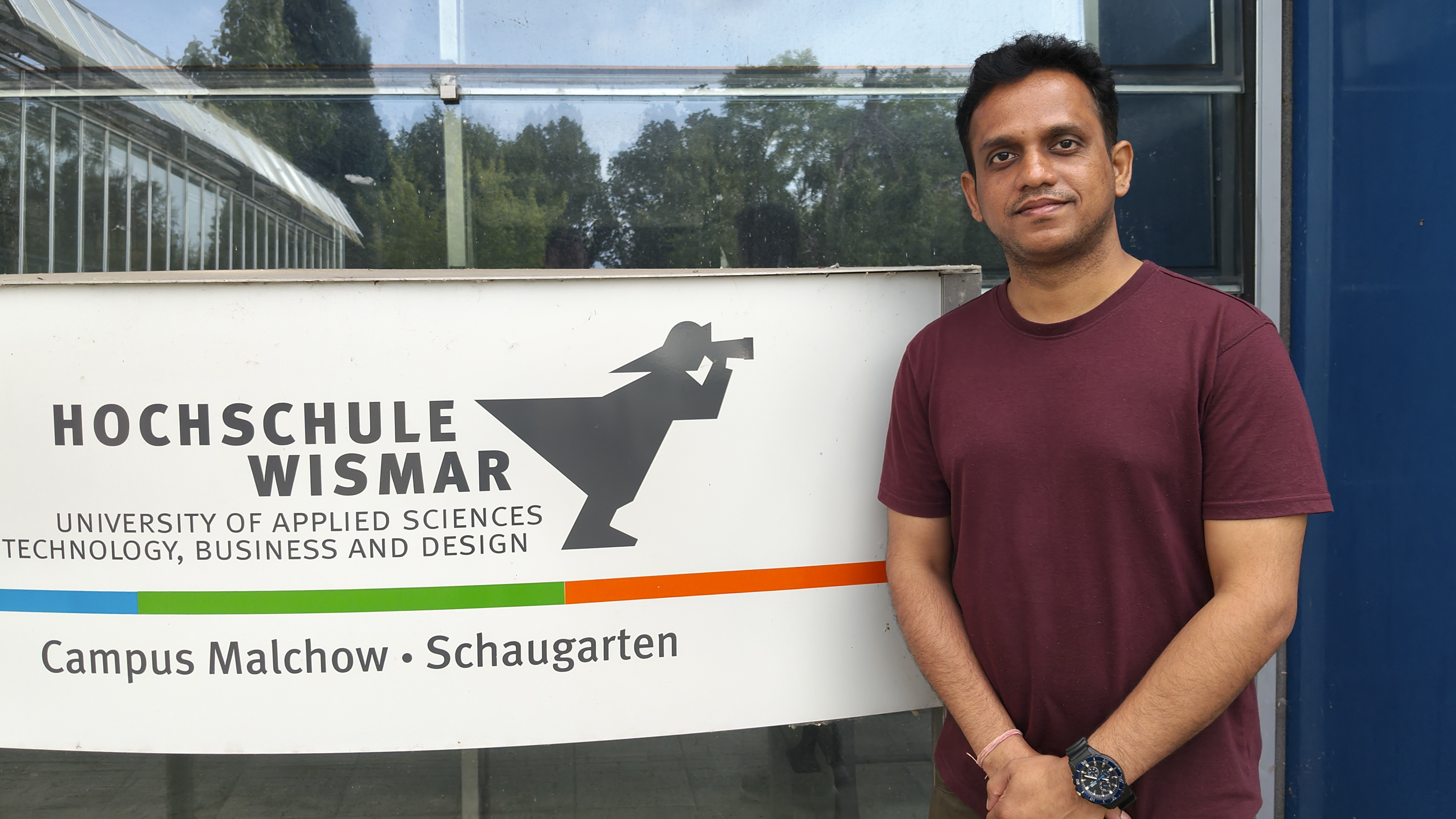 Ein Mann steht vor einem Schild der Hochschule Wismar am Campus Malchow, das den Schriftzug „HOCHSCHULE WISMAR – University of Applied Sciences: Technology, Business and Design“ sowie das Logo der Hochschule zeigt. Im Hintergrund sind Glasfassaden und Bäume zu erkennen.