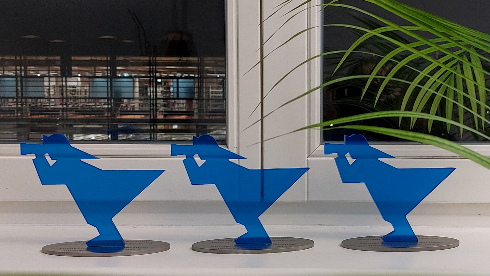 Die drei blau gefärbten Pokale in Form des Fischer-Signets aus dem Hochschul-Logo stehen auf einem Fensterbrett. Durch das Fesnter hinweg kann man den dunklen Himmel und die erleuchtete Bibliothek sehen.