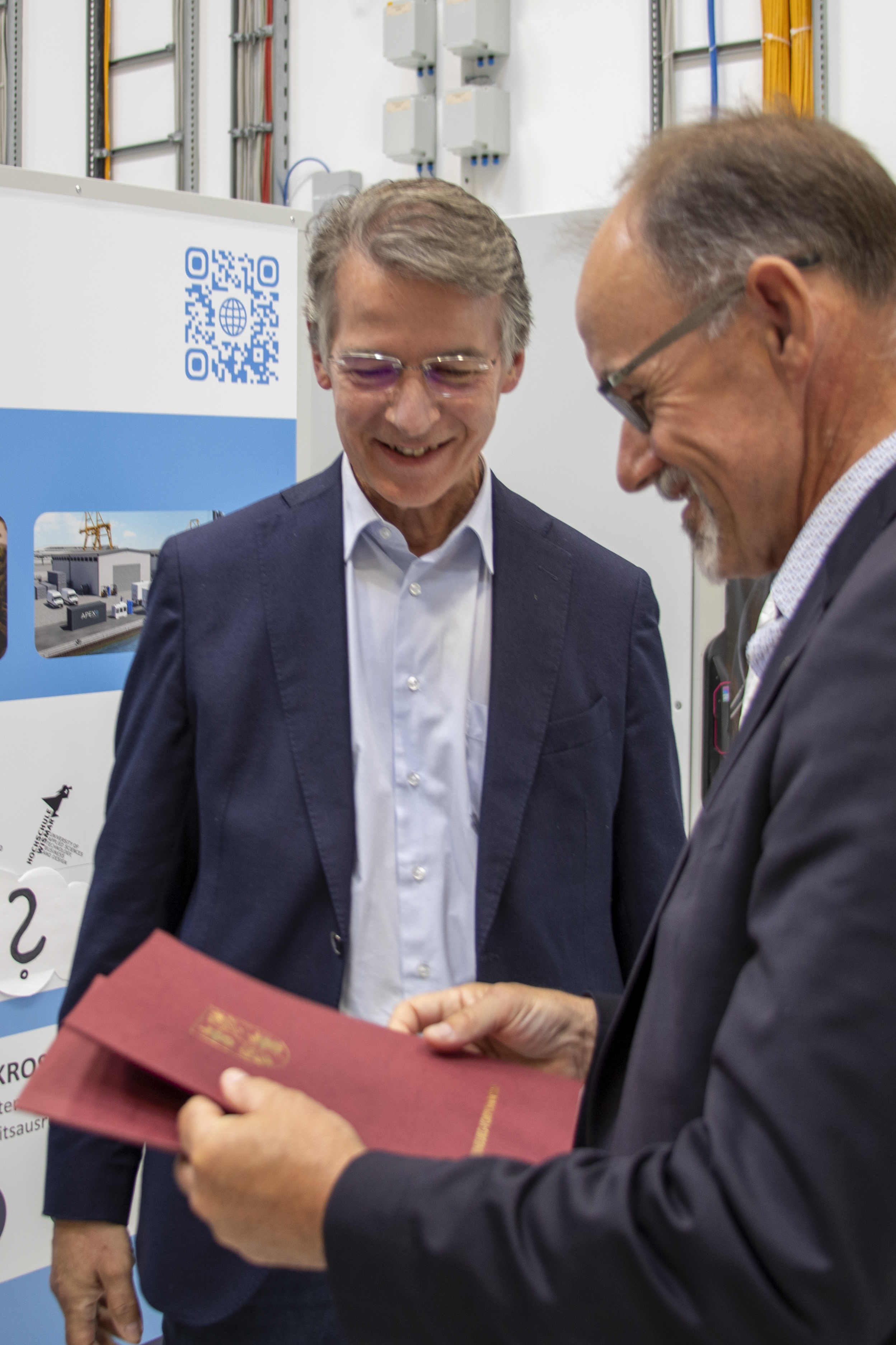 Der Minister ist von der Seite und der Professor von vorn zu shen. Beide schauen auf eine rote Mappe, die sie in den Händen halten.