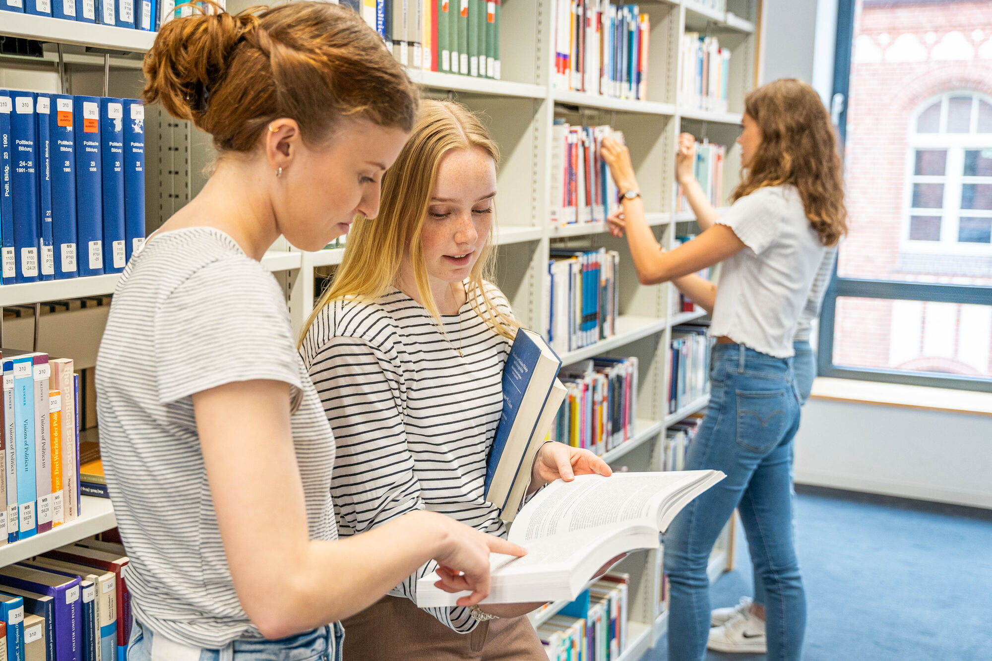 Drei junge Frauen stehen in einer Bibliothek vor Bücherregalen. Zwei von ihnen schauen gemeinsam in ein aufgeschlagenes Buch, während die dritte ein Buch aus dem Regal nimmt.
