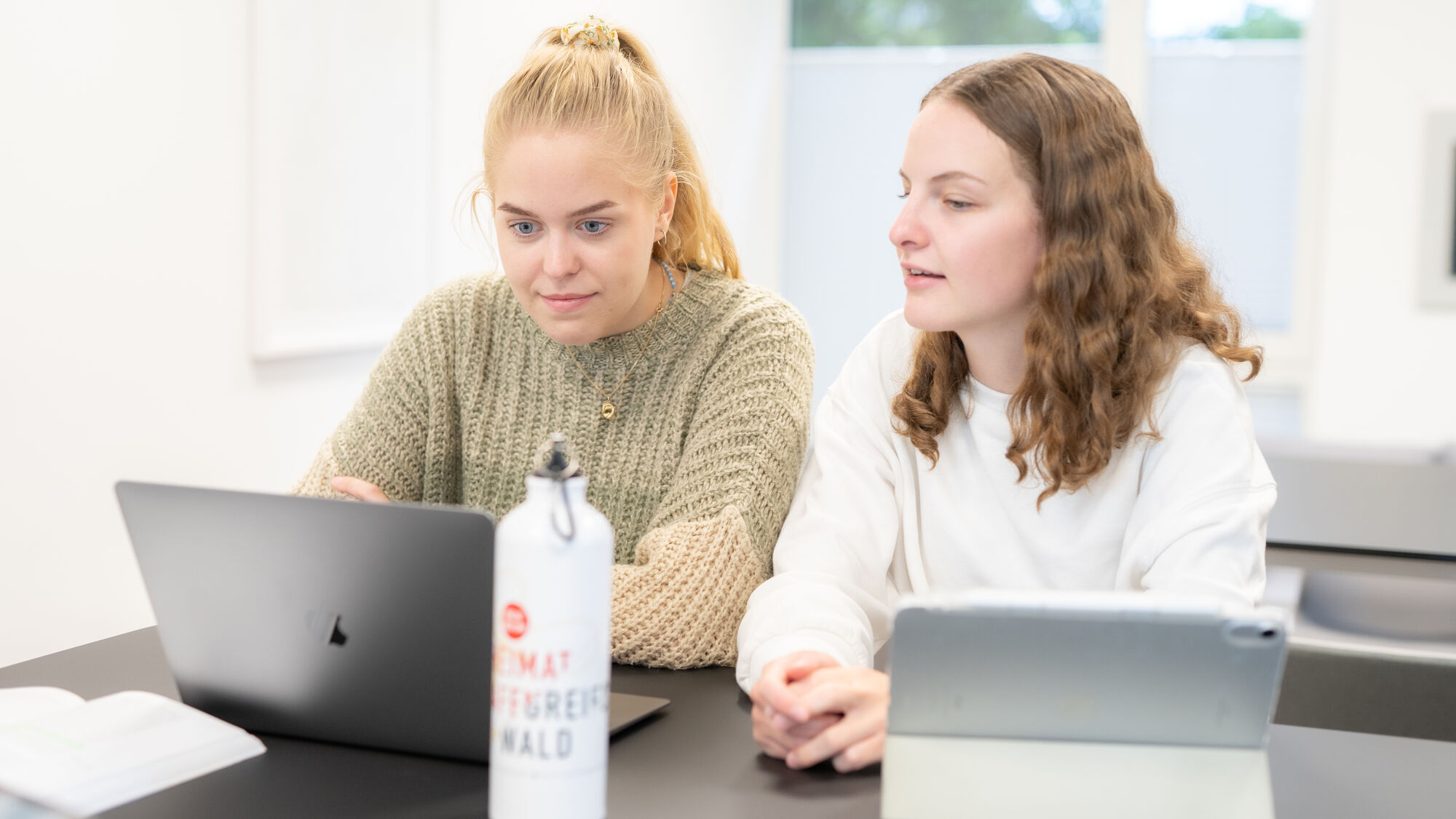 Zwei Studentinnen sitzen nebeneinander an ihren Notebooks und tauschen sich aus.