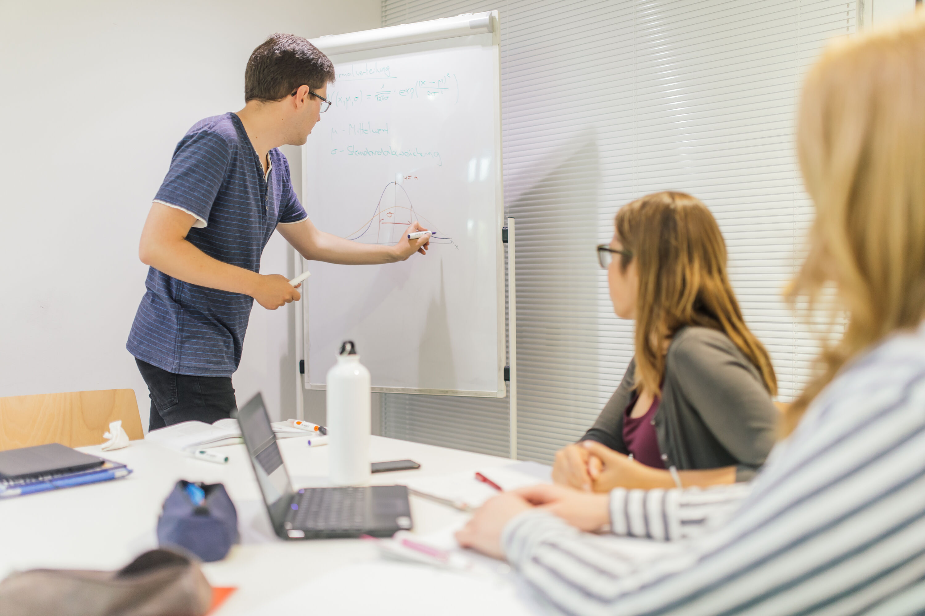 Symbolbild Studium Eine männliche Person schreibt etwas auf ein Whiteboard, während zwei weibliche Personen zuschauen. Seminarsituation