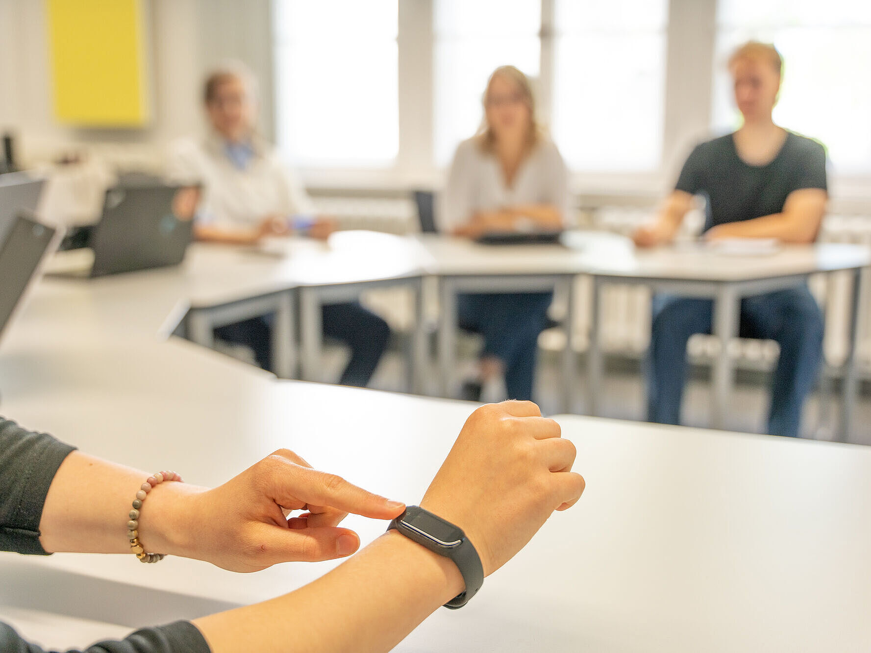 Symbolbild Meeting Im Vordergrund wird eine Smartwatch an einem Handgelenk bedient, während im Hintergrund unscharf vier Personen an Tischen in einem hellen Raum sitzen.
