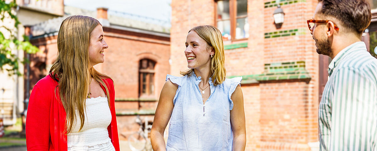 Eine Gruppe von zwei Studentinnen und einem Student stehen bei Sonnenschein vor dem Backsteingebäude der Germanistik und unterhalten sich.