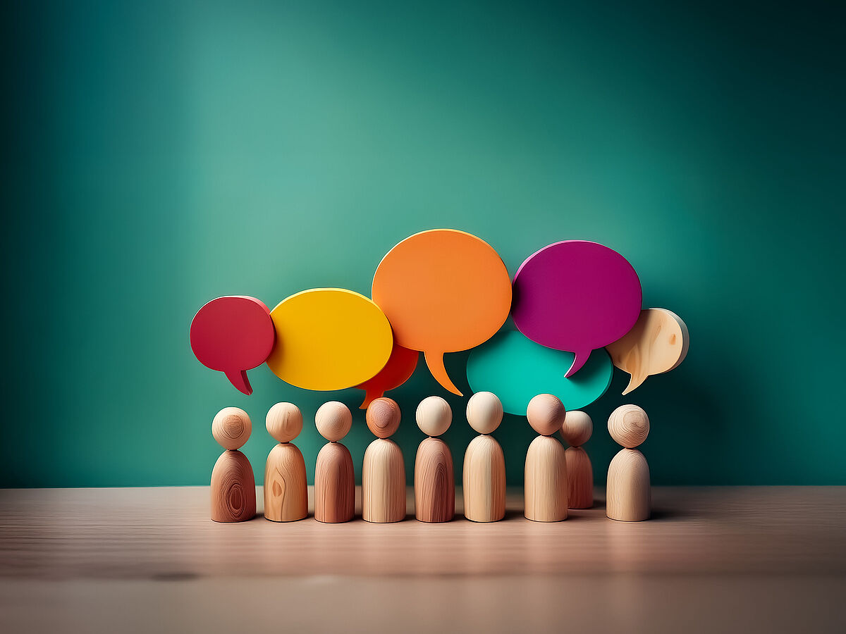 Symbolbild Dialog Vor einer grünen Wand stehen aufgereiht auf einem Holztisch kleine Holzfiguren, hinter ihnen sind bunte Sprechblasen an der Wand zu sehen.