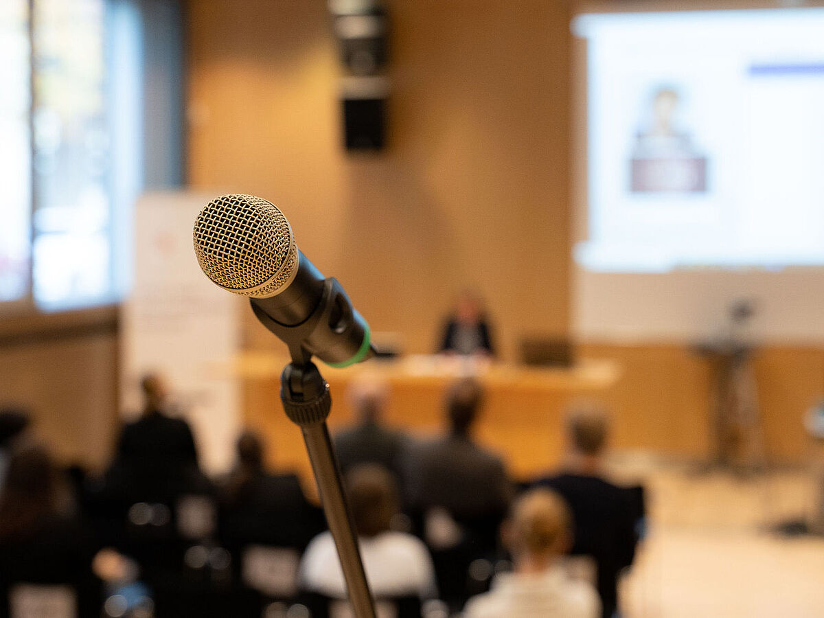 Symbolbild akademische Karriere Ein Mikrofon im Vordergrund, scharfgestellt, mit einem unscharfen Konferenzraum im Hintergrund, in dem Personen einer Präsentation folgen.