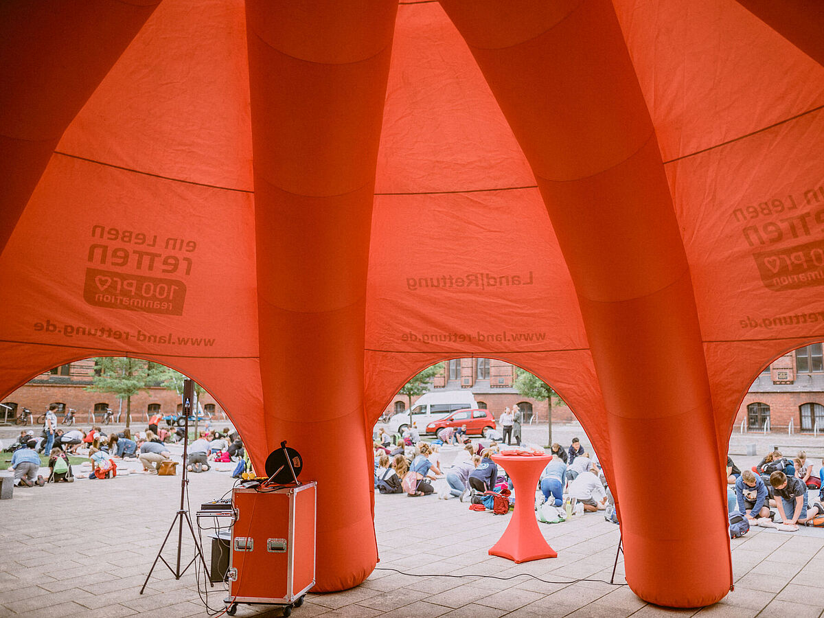 Zu sehen ist ein großes orangefarbenes Zelt. Im Hintergrund knien viele Kinder während einer Rettungsübung zur Wiederbelebung von Menschen.