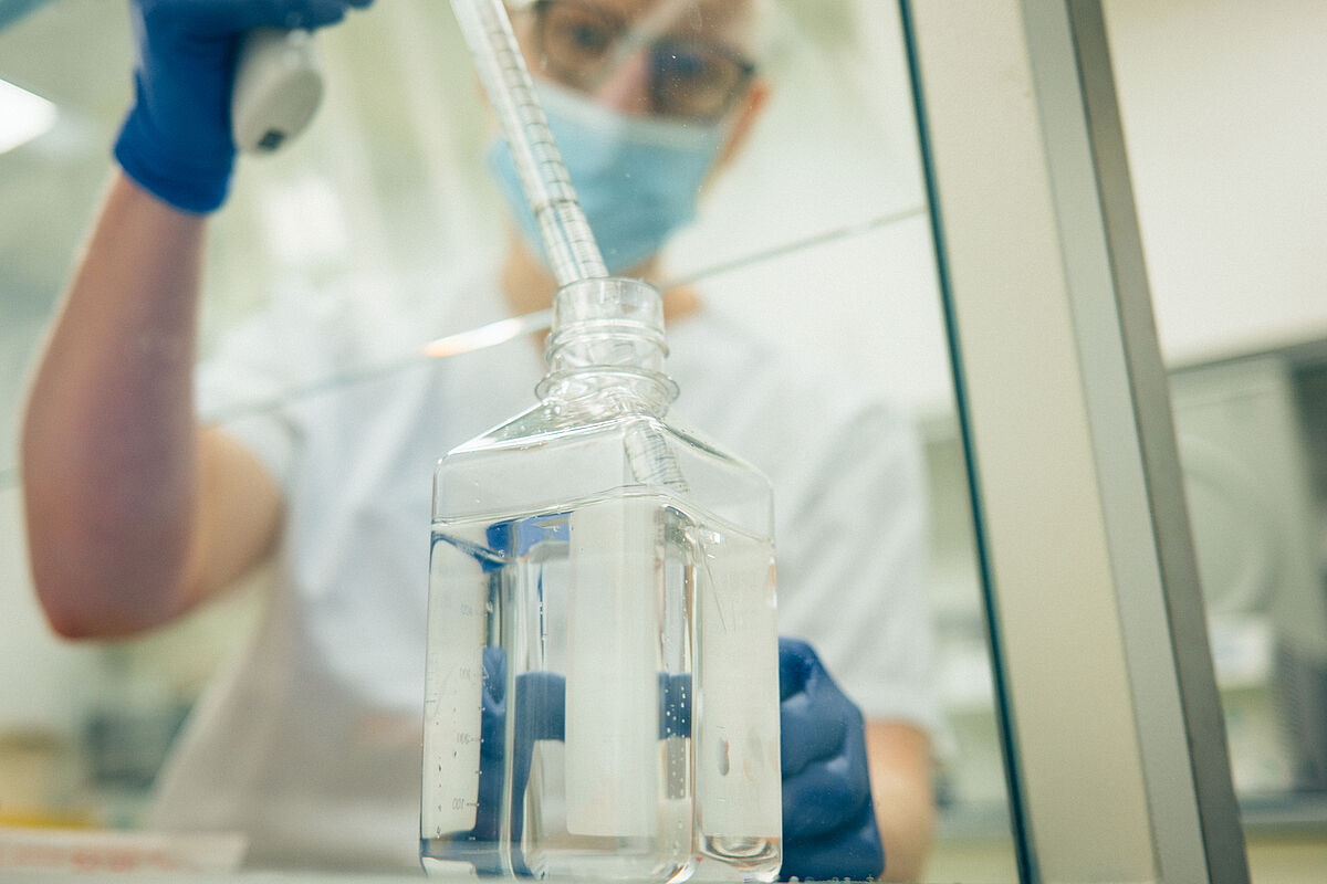 Eine verschwommen dargestellte Person mit blauem Handschuh entnimmt mit einer Pipette eine durchsichtige Flüssigkeit aus einem Glasbehälter. 