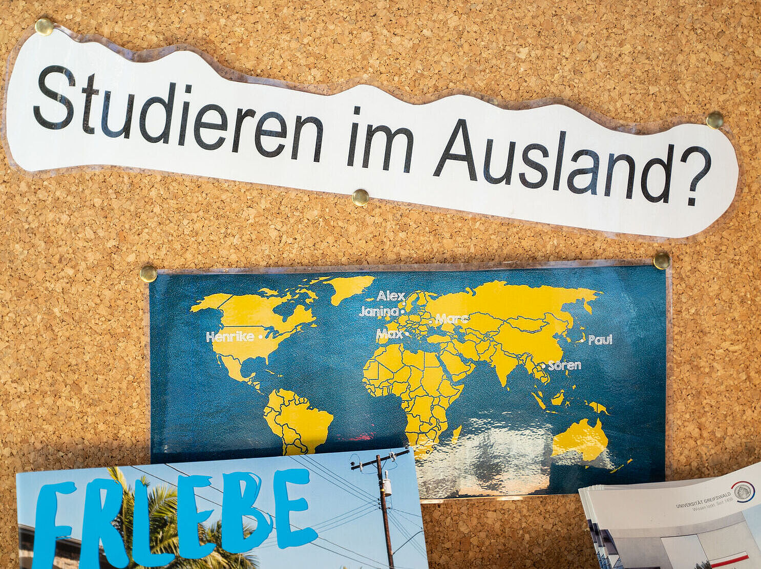 Eine Pinnwand mit der Überschrift „Studieren im Ausland?“ und einer Weltkarte, auf der Namen markiert sind, darunter Flyer im unteren Bereich.