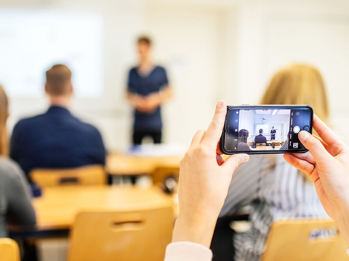 Im Vordergrund hält eine Person ein Smartphone in der Hand, im Hintergrund sind weitere Personen sehr verschwommen zu sehen.