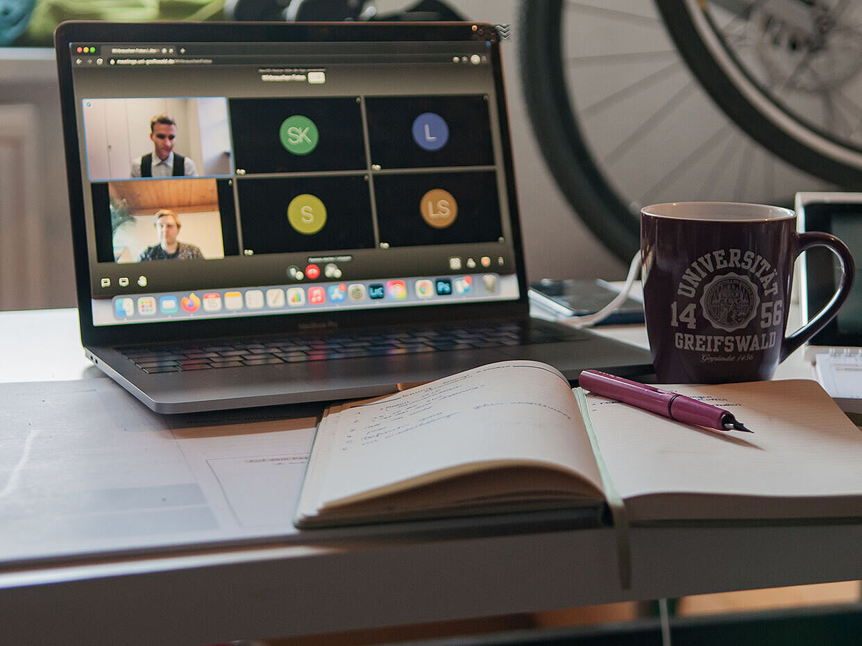 Auf einem Schreibtisch steht ein offenes Notebook, auf dem eine Videokonferenz läuft. Daneben steht eine Tasse und ein aufgeschlagenes Buch mit einem Stift liegen davor.