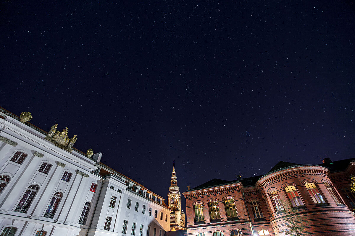 Auf dem Bild ist der Historischer Campus mit Sternenhimmel zu sehen. Links erkennt man das hauptgebäude und Rechts das Alte Audimax. Dazwischen ist der beleuchtete Mond zu sehen.