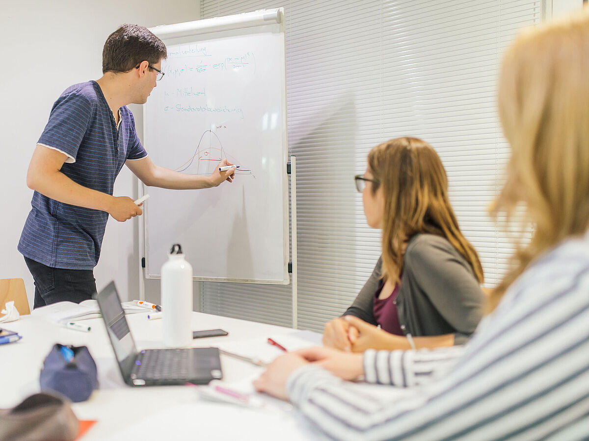 Eine männliche Person schreibt etwas auf ein Whiteboard, während zwei weibliche Personen zuschauen. Seminarsituation