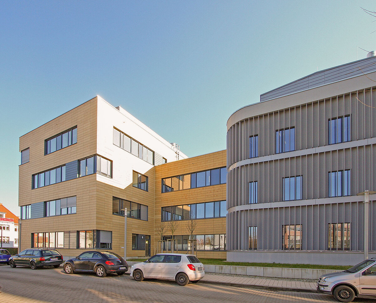 Symbolbild Gebäude C_DAT Gebäude C_DAT: Das moderne Gebäude mit rechteckigen und geschwungenen Fassaden aus Holz und Metalllamellen. Davor stehen geparkte Autos auf einer gepflasterten Straße.