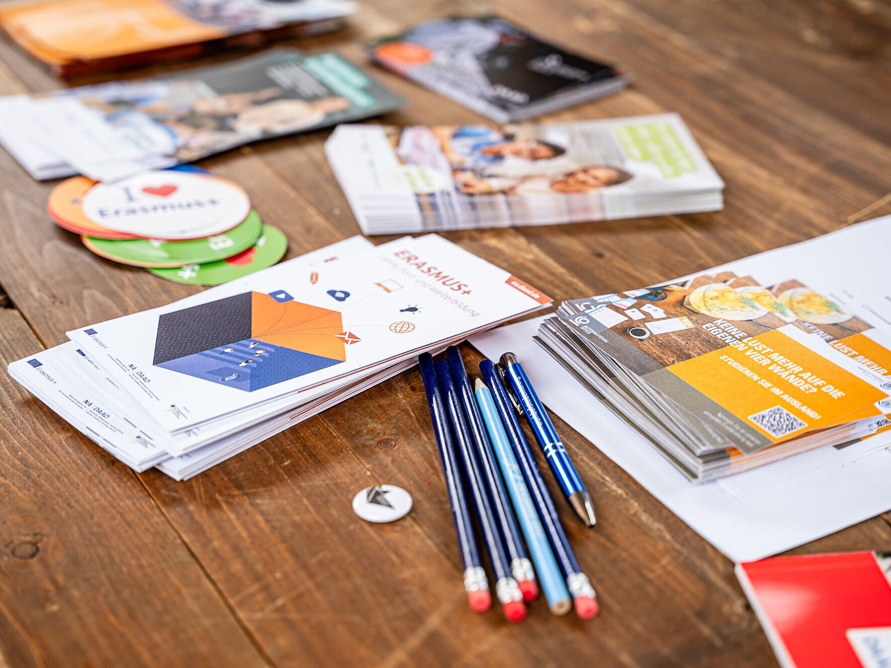 Nahaufnahme von Informationsmaterialien für Erasmus+ auf einem Tisch. Im Vordergrund sind Stifte und Faltblätter zu sehen.
