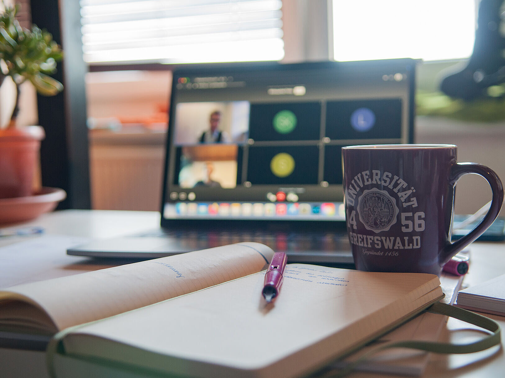 Ein Schreibtisch mit einem aufgeklappten Laptop, auf dem ein Videocall läuft, einer Tasse mit Universitätslogo und einem offenen Notizbuch mit Stift.