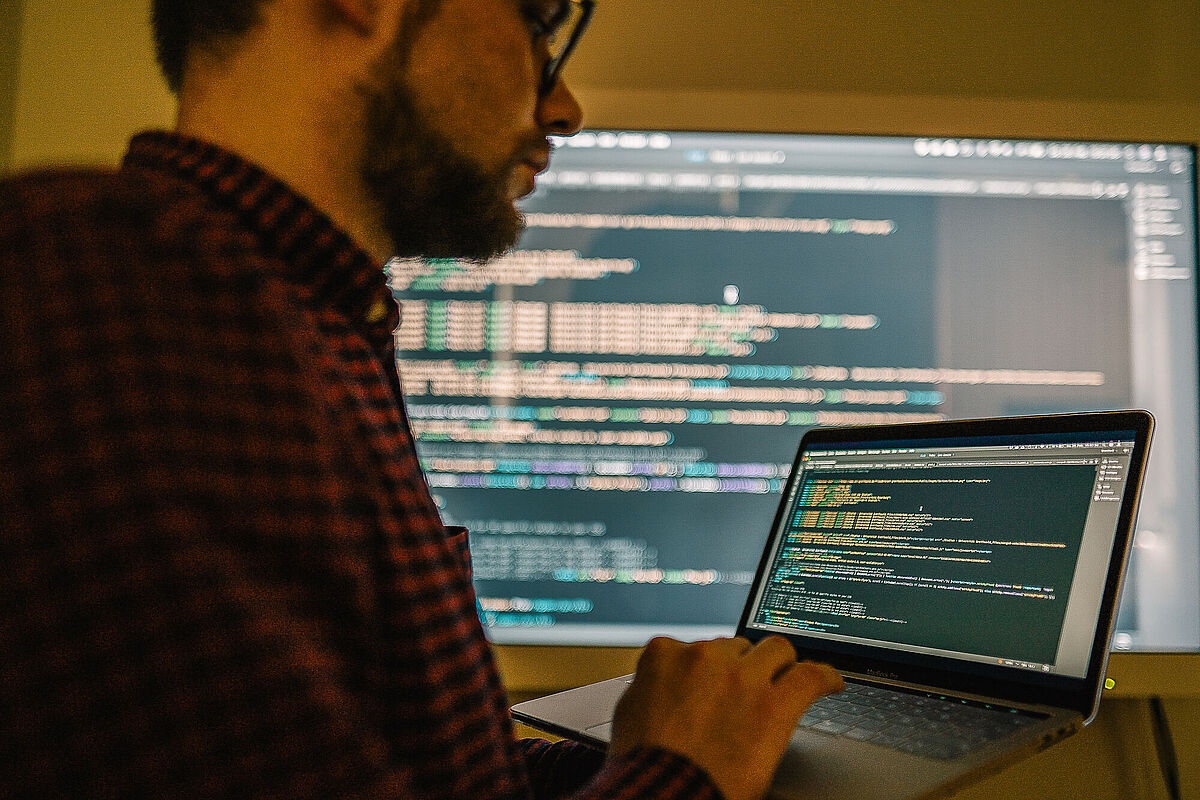 Ein Mann bearbeitet auf einem Notebook einen Programmiercode, der auf einem beleuchteten Bildschirm ausgespielt wird.