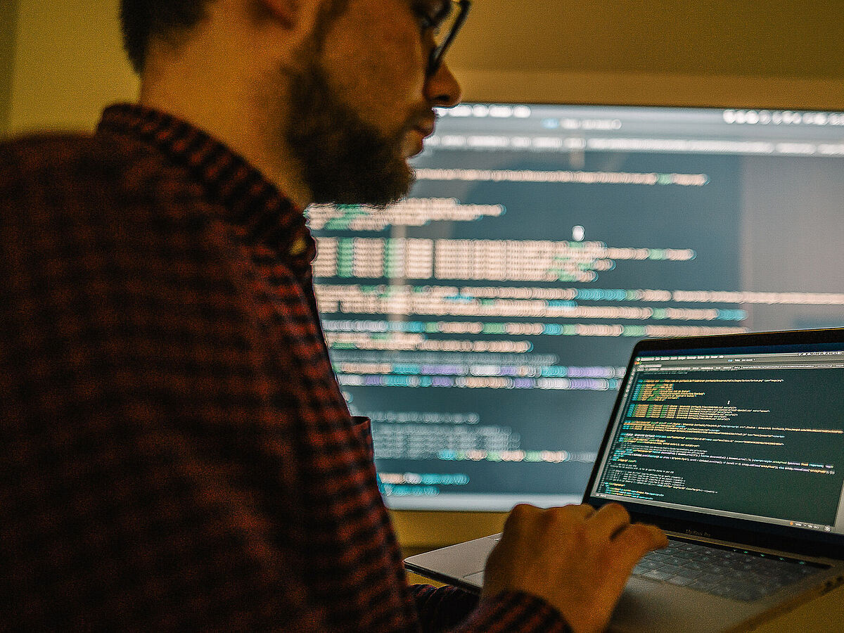 Ein Mann bearbeitet auf einem Notebook einen Programmiercode, der auf einem beleuchteten Bildschirm ausgespielt wird.
