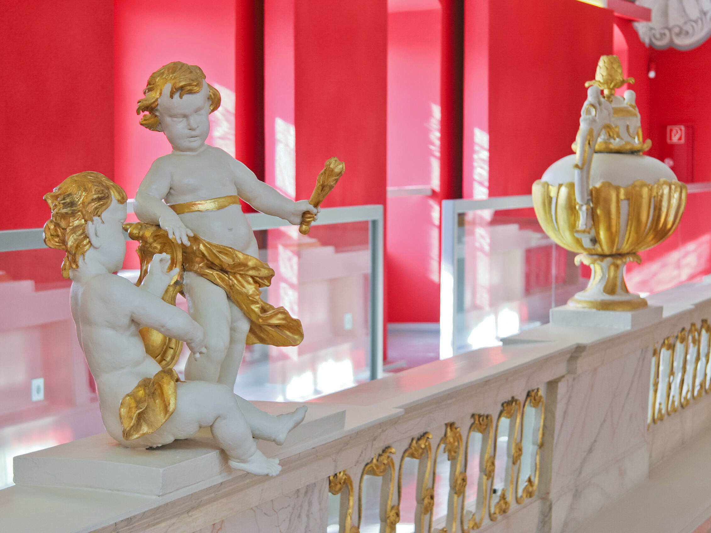 Vor den roten Wänden der Uni-Aula sind Figuren, sog. Putten, zu sehen. Sie sind aus weißem Marmor und mit Gold abgesetzt.