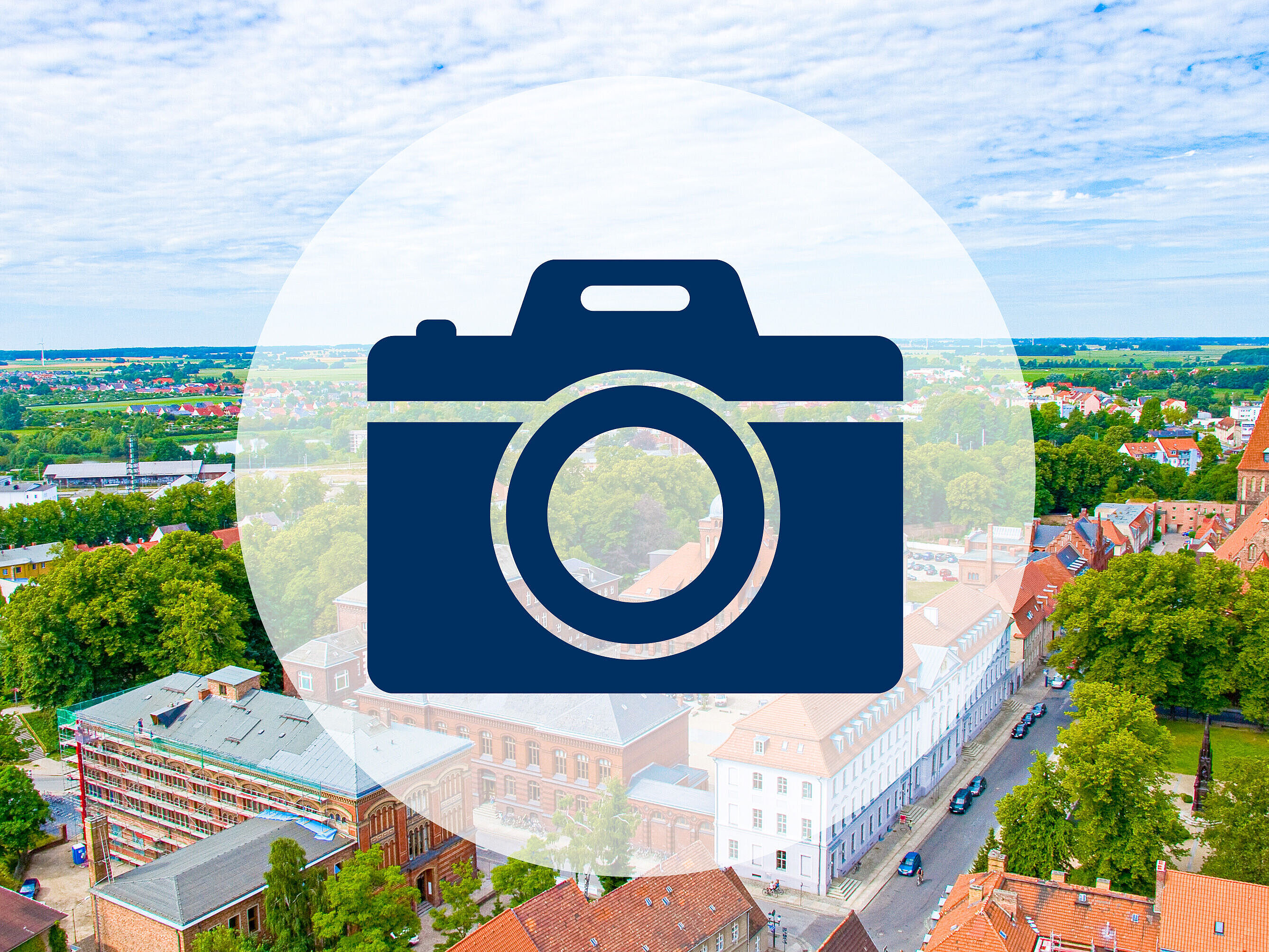 Auf einer freundlich hellen Luftaufnahme des historischen Campus der Uni Greifswald ist ein blaues Icon einer Fotokamera.