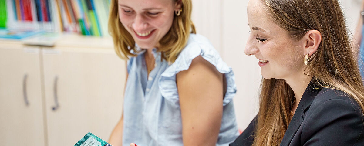 Zwei weibliche Studierende schauen sich gemeinsam ein Buch an und freuen sind dabei.