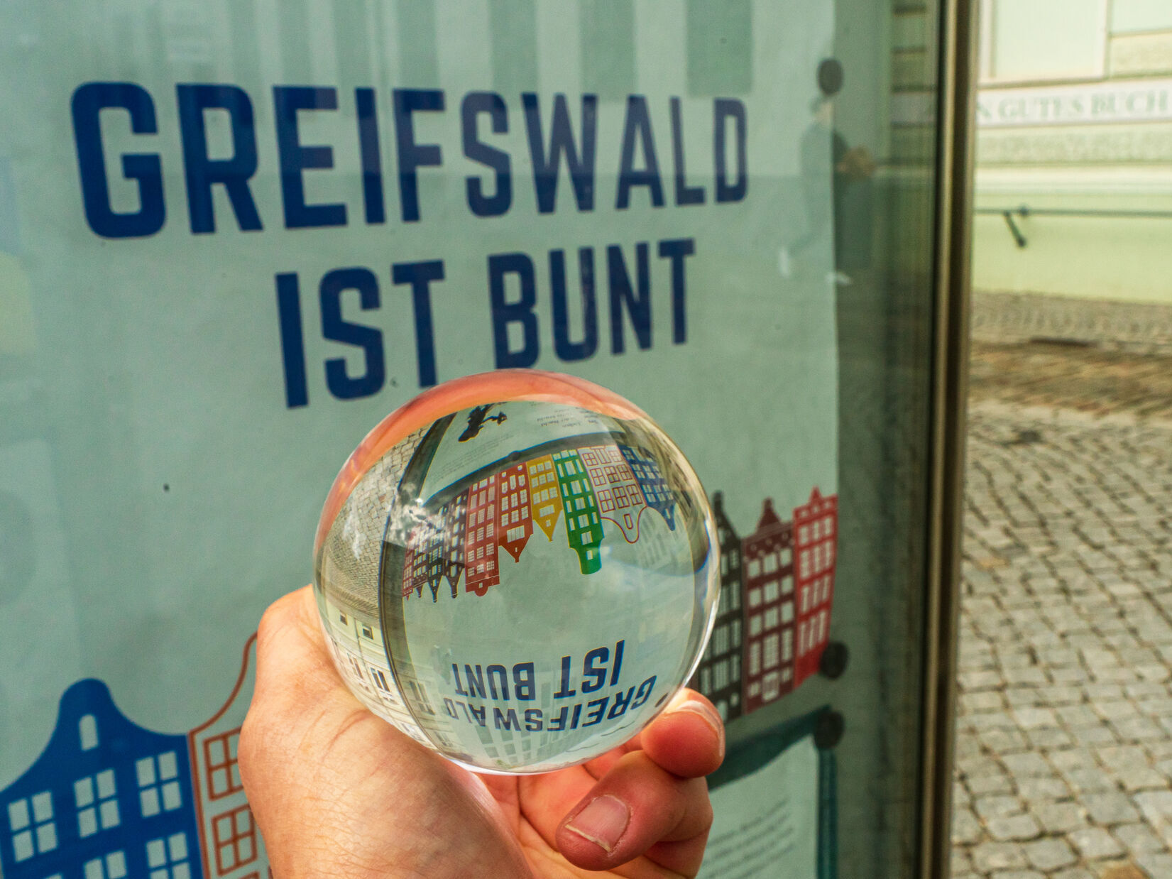 Eine Hand hält eine Glaskugel vor einem Plakat mit der Aufschrift „Greifswald ist bunt“.